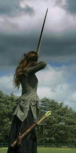 A woman in medieval costume faces away from the viewer, pointing a longbow.
