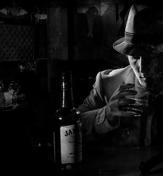 A black and white photo of a man in a fedora hunched over a bottle of whiskey, smoking a cigarette.