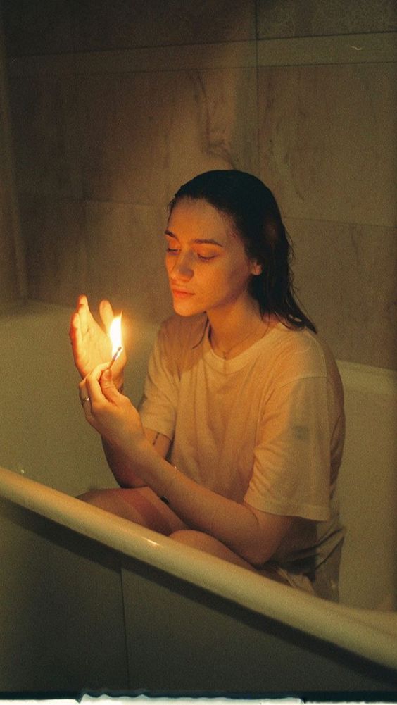 A girl sits in a bathtub in a white tshirt, contemplating a lit match