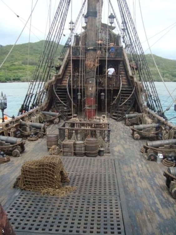 Image of a tall ship taken from onboard the deck, looking back towards the poop. Rigging, cannons, barrels are visible.