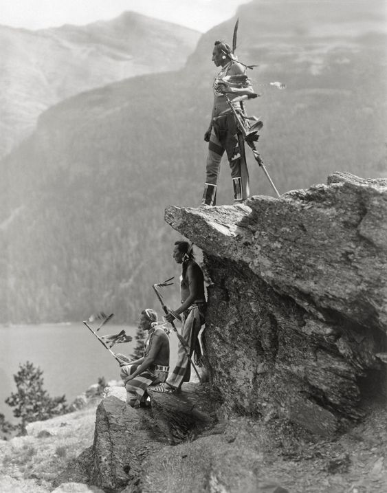 Three Blackfoot tribe members in full battle regalia stand on a rocky cliff. Black and white photograph, circa 1910.